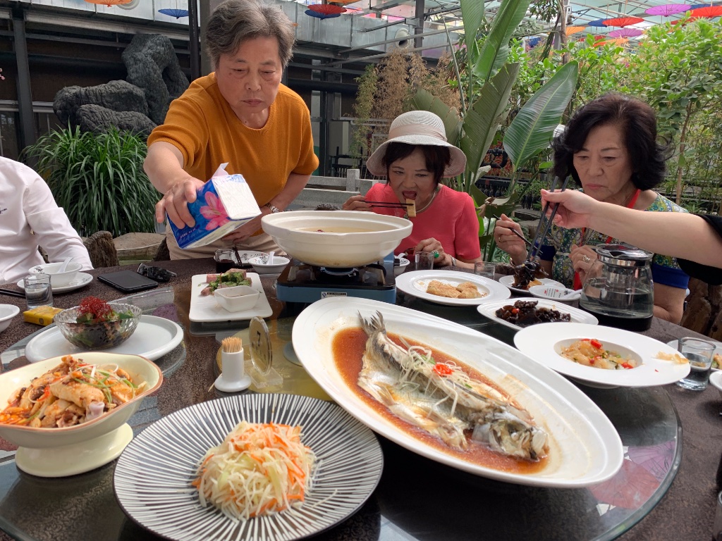 Hier durften wir an großen runden Tischen zwischen Pflanzen und Wasserläufen fürstlich tafeln. Die Dame im orangen Pullover, die gerade nach der Schachtel mit den Papierservietten greift, ist die Präsidentin der chinesischen Frauen-Penjing-Vereinigung. Leider weiß ich ihren Namen nicht mehr